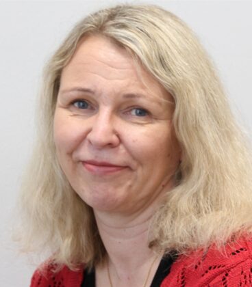 Hartioihin ulottuvat vaaleat hiukset ja punainen toppi hymyilee pehmeästi yksinkertaista taustaa vasten.