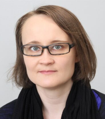 Lyhyet, vaaleanruskeat hiukset ja lasit omaava henkilö on yllään tumma toppi ja musta huivi. Tausta on neutraali ja vaaleanharmaa.