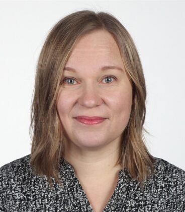 Henkilö, jolla on olkapäille ulottuvat vaaleanruskeat hiukset, hymyilee hieman. Heillä on yllään mustavalkokuvioinen toppi. Tausta on tavallinen valkoinen.