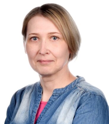 Lyhyet vaaleat hiukset omaava nainen, jolla on farkkupaita ja vaaleanpunainen toppi, katsoo kameraa neutraalilla ilmeellä valkoista taustaa vasten.