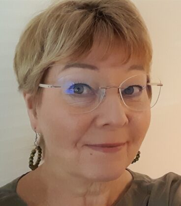 Lyhythiuksinen vaaleanruskea tukka ja silmälasit käyttävä nainen hymyilee kameralle. Hänellä on vihreät korvakorut ja vaalea toppi, ja hänen takanaan on neutraali tausta.