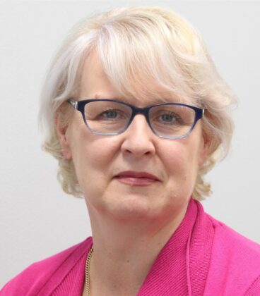 Henkilö, jolla on lyhyet, vaaleat hiukset ja silmälasit, katsoo kameraan. He ovat pukeutuneet kirkkaan vaaleanpunaiseen toppiin ja kultaiseen kaulakoruun. Tausta on tasainen ja valkoinen.