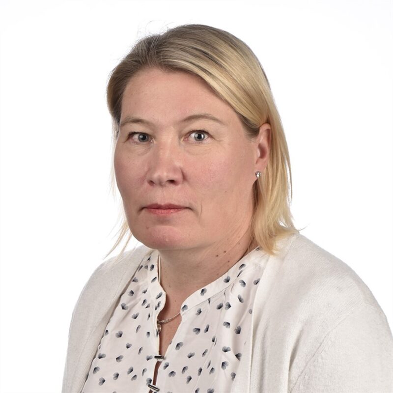 Hartioihin ulottuvat vaaleat hiukset omaavalla henkilöllä on yllään valkoinen, pienikuvioinen pusero ja vaalea neuletakki. he poseeraavat tavallista valkoista taustaa vasten.