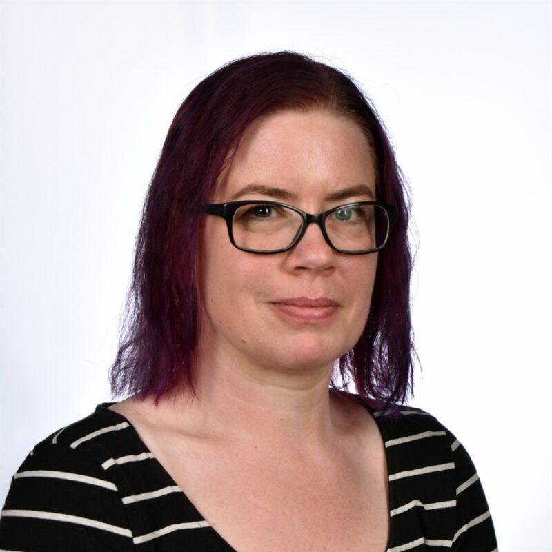 Henkilö, jolla on olkapäille ulottuvat violetit hiukset ja silmälasit, yllään musta paita, jossa on valkoiset raidat, katsoo kameraa valkoista taustaa vasten.