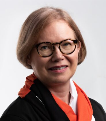 Hymyilevä lyhyet vaaleanruskeat hiukset omaava nainen, jolla on silmälasit, musta asu ja oranssi huivi valkoisen paidan päällä, yksiväristä vaaleaa taustaa vasten.