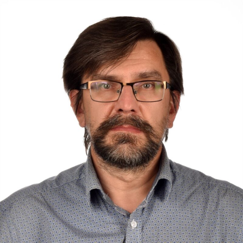 Miehellä, jolla on tummat hiukset, silmälasit ja parta, on yllään kuviollinen paita ja hän katsoo suoraan kameraan valkoista taustaa vasten.