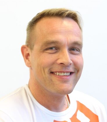 Mies, jolla on lyhyet vaaleat hiukset, hymyilee kameralle. Hänellä on yllään valkoinen t-paita, jossa on oransseja aksentteja, ja hän poseeraa valkoista taustaa vasten.