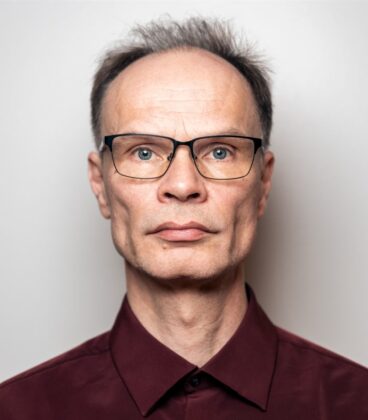 Henkilöllä, jolla on lyhyet hiukset ja silmälasit, on yllään kastanjanruskea napillinen paita. He seisovat valkoista taustaa vasten ja katsovat suoraan kameraan neutraalilla ilmeellä.