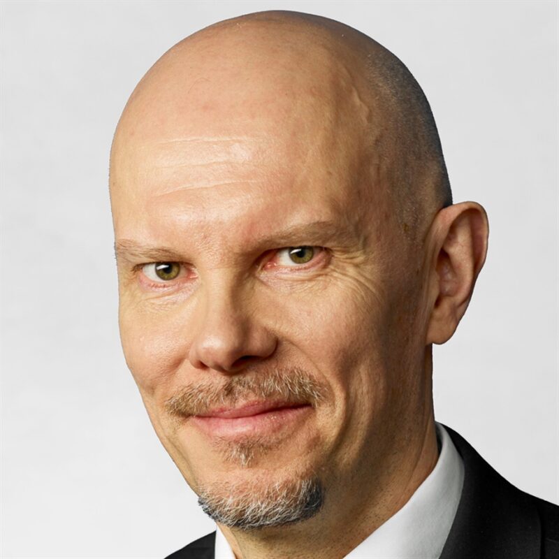 Kalju mies, jolla on vaalea pukkiparta, hymyilee hieman. Hänellä on yllään musta puku ja valkoinen paita ja hän näyttää olevan vaaleanharmaalla taustalla.