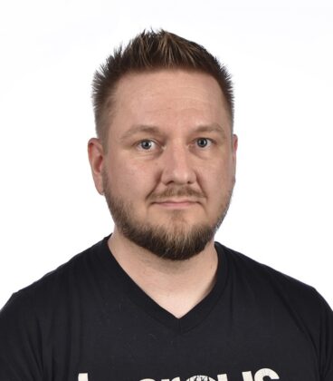 Lyhyt, piikikäs tukka ja siisti partainen mies, jolla on musta t-paita, katsoo suoraan kameraan neutraalilla ilmeellä yksinkertaista valkoista taustaa vasten.