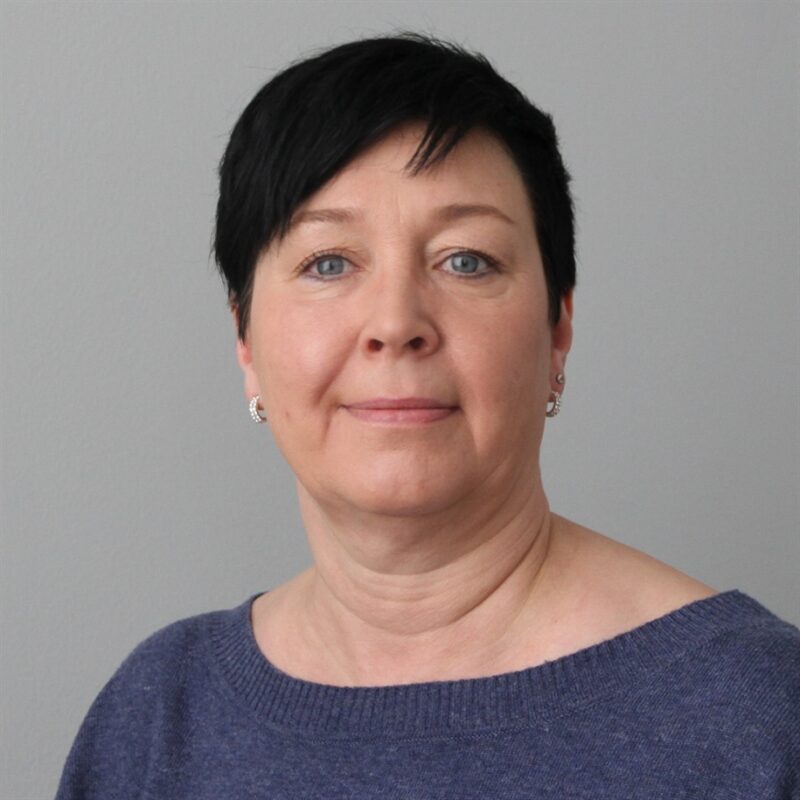 Lyhyet mustat hiukset omaavalla henkilöllä on yllään sininen pusero ja pienet korvakorut. He poseeraavat tavallisen harmaan taustan edessä.