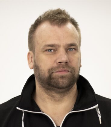 Mies, jolla on lyhyet hiukset ja parta yllään tumma villapaita, katsoen suoraan kameraan tavallista valkoista taustaa vasten.
