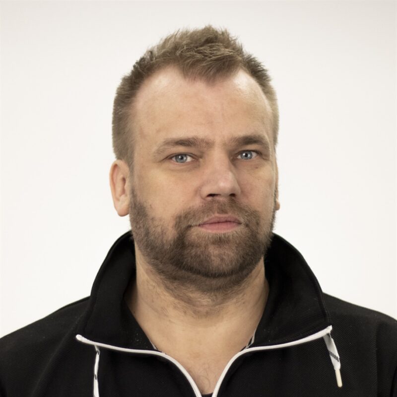 Mies, jolla on lyhyet hiukset ja parta yllään tumma villapaita, katsoen suoraan kameraan tavallista valkoista taustaa vasten.