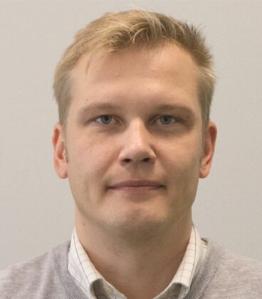 Lyhythiuksinen mies, jolla on vaaleanharmaa neulepusero valkokauluspaidan päällä, katsoo suoraan kameraan neutraalilla ilmeellä yksinkertaista, vaaleaa taustaa vasten.