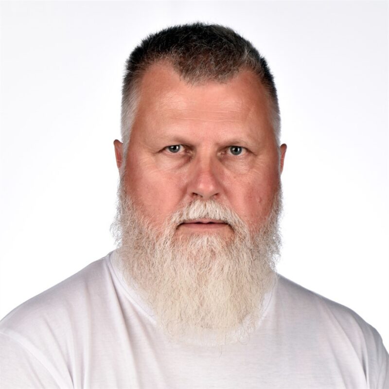 Keski-ikäisellä miehellä, jolla on valkoinen parta ja lyhyet hiukset, on päällään tavallinen valkoinen paita ja hän katsoo suoraan kameraan valkoista taustaa vasten.