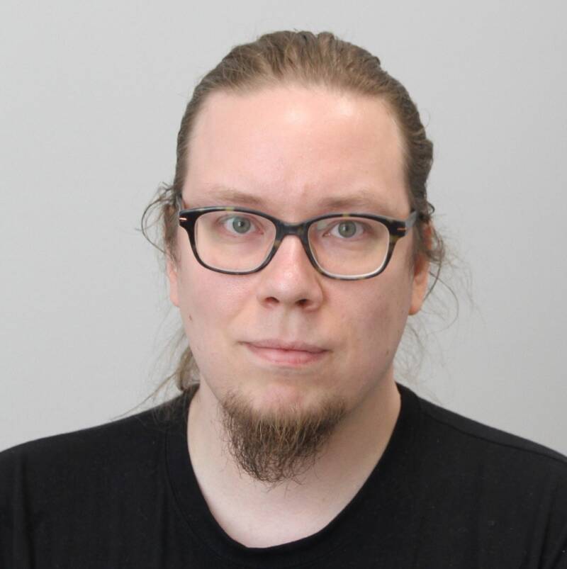 Pitkät hiukset taakse sidottu henkilö, jolla on mustareunaiset lasit ja musta paita, katsoo suoraan kameraan vaaleaa taustaa vasten. Heillä on lyhyt parta ja viikset.
