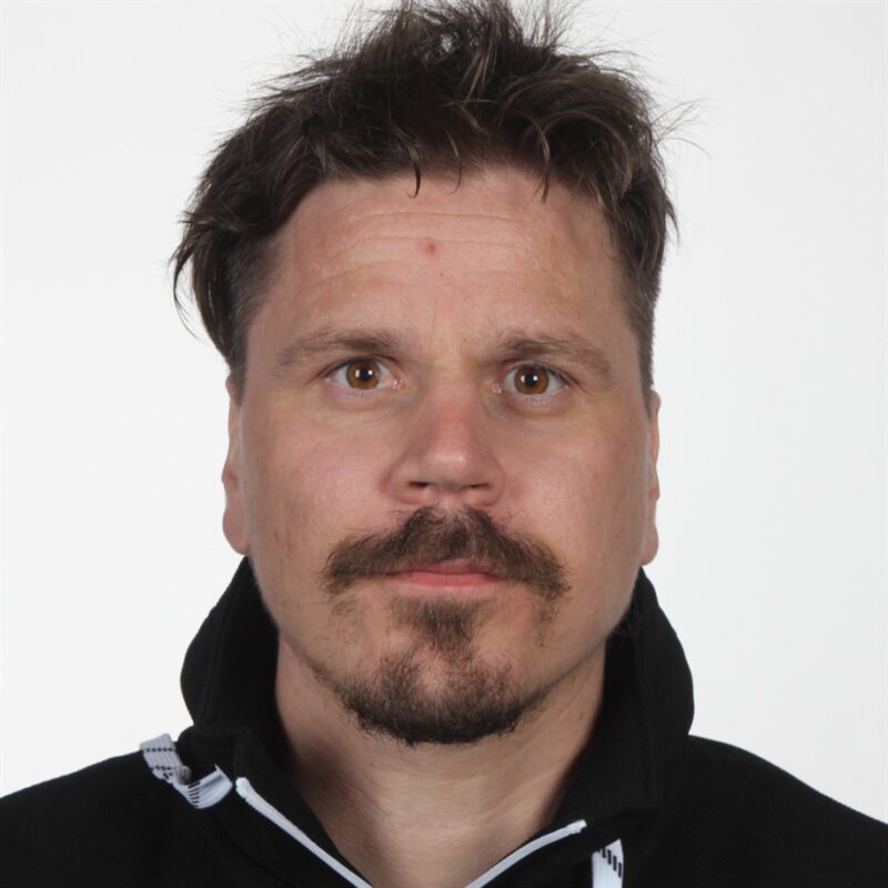 Henkilö, jolla on lyhyet ruskeat hiukset, viikset ja vuohipaita, jolla on musta vetoketjullinen takki. He katsovat suoraan kameraan neutraalilla ilmeellä. Tausta on tavallinen valkoinen.