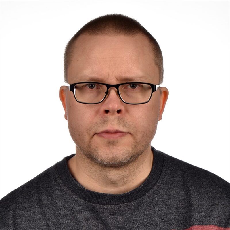Henkilö, jolla on lyhyet hiukset ja silmälasit, tuijottaa suoraan kameraan. Heillä on yllään tumma paita ja tausta on pelkkä valkoinen.