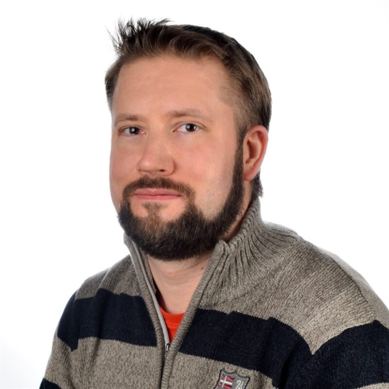 Miehellä, jolla on lyhyet hiukset ja parta, on yllään harmaa pusero, jossa on mustat raidat ja merkki. Tausta on tavallinen valkoinen.