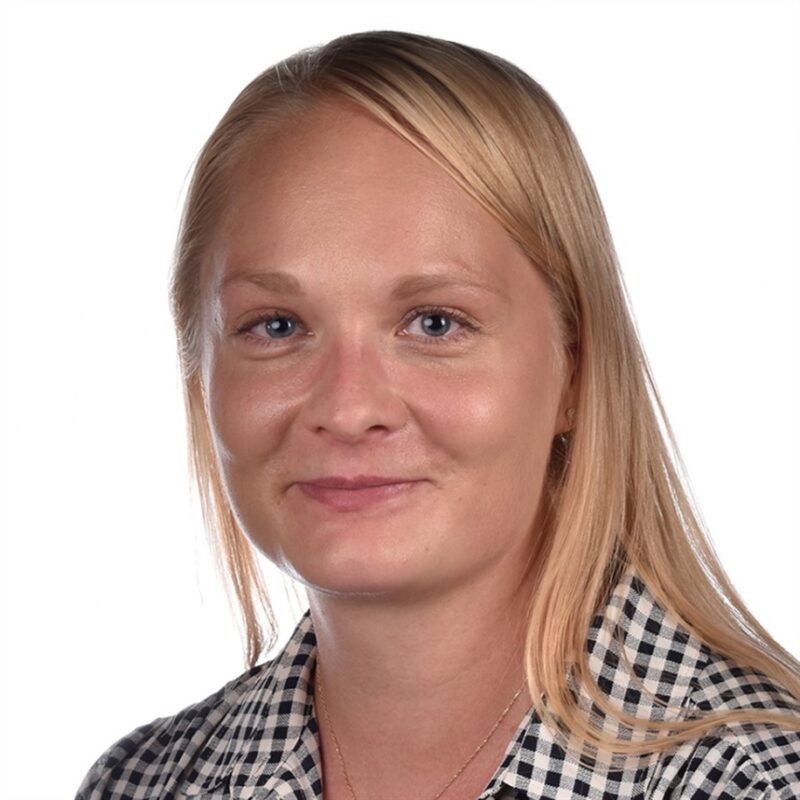 Nainen, jolla on pitkät vaaleat hiukset ja yllään mustavalkoinen ruudullinen paita, hymyilee neutraalisti. Tausta on tasainen ja valkoinen.