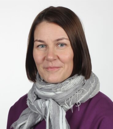 Suorat ruskeat hiukset omaava nainen, jolla on violetti toppi ja vaaleanharmaa huivi, hymyilee pehmeästi tavallisen valkoisen taustan edessä.