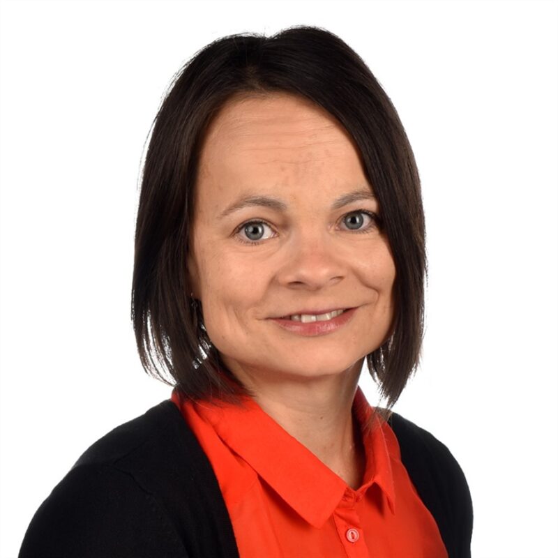 Mies, jolla on olkapäille ulottuvat ruskeat hiukset, hymyilee kameralle. Heillä on yllään punainen pusero ja musta neuletakki, jotka on asetettu valkoista taustaa vasten.