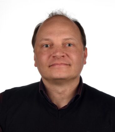 Lyhythiuksinen henkilö, jolla on tumma villapaita kauluspaidan päällä, hymyilee kameralle valkoista taustaa vasten.