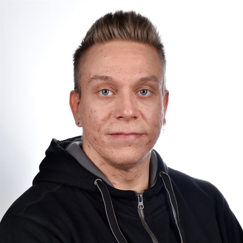 Lyhyet, piikkihiuksinen ja mustaa hupparia pukeutunut henkilö katsoo suoraan kameraan valkoista taustaa vasten.