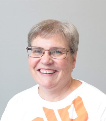 Hymyilevä, lyhyet harmaat hiukset ja silmälasit omaava henkilö, jolla on valkoinen, oranssikuvioinen paita, poseeraa vaaleanharmaan taustan edessä.