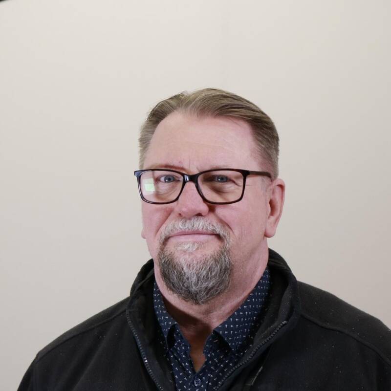 Keski-ikäinen mies, jolla on silmälasit, lyhyet vaaleanruskeat hiukset ja vuohenparkki hymyilee hieman. Hänellä on yllään tumma takki laivastonsinisen paidan päällä, jossa on pieniä valkoisia pilkkuja. Tausta on tasainen ja vaalea.