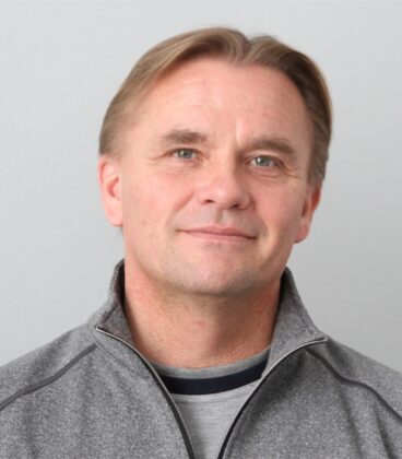 Lyhyet, vaaleanruskeat hiukset omaavalla henkilöllä on yllään harmaa vetoketjullinen pusero ja musta paita. Niissä on neutraali ilme ja niiden takana on yksinkertainen vaaleanharmaa tausta.