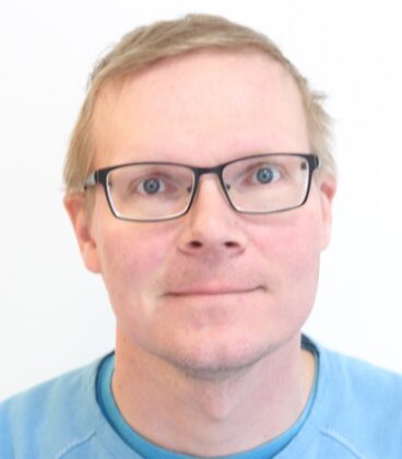 Henkilö, jolla on lyhyet, vaaleat hiukset ja silmälasit, katsoo hieman ylöspäin. Heillä on yllään vaaleansininen paita ja he ovat puhtaan valkoisen taustan edessä.