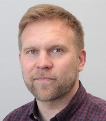 Henkilö, jolla on lyhyet, vaaleanruskeat hiukset ja parta, katsoo kameraan. Heillä on yllään punainen ja musta ruudullinen paita ja he seisovat tavallista vaaleaa taustaa vasten.