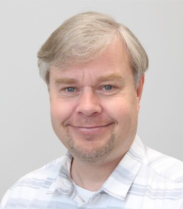 Keski-ikäinen vaaleahiuksinen ja pukkiparta hymyilee kameralle. Heillä on yllään valkoinen ja harmaa raidallinen paita tavallista taustaa vasten.
