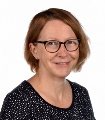Nainen, jolla on lyhyet, vaaleanruskeat hiukset ja silmälasit, hymyilee kameralle. Hänellä on yllään musta toppi, jossa on valkoisia pilkkuja. Tausta on tavallinen valkoinen.