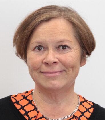 Keski-ikäinen lyhyttukkainen vaaleanruskeahiuksinen nainen hymyilee kameralle. Hänellä on yllään musta paita, jossa on oranssikuvioinen kaulus ja hopeinen ketjukaulakoru. Kuva on poseerattu yksinkertaista valkoista taustaa vasten.