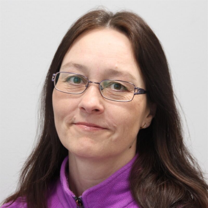 Pitkät ruskeat hiukset ja lasit omaava henkilö on pukeutunut violettiin toppiin ja hymyilee lempeästi yksinkertaista taustaa vasten.