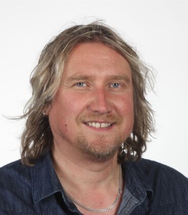 Hymyilevä yksilö, jolla on pitkät, aaltoilevat hiukset ja parta, yllään tumma denimpaita ja hopeakaulakoru, valkoista taustaa vasten.
