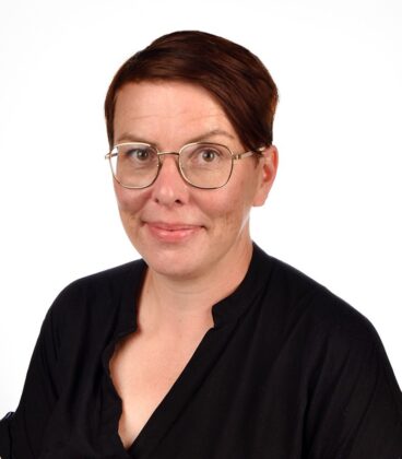 Henkilö, jolla on silmälasit ja musta toppi, hymyilee kameralle. Heidän hiuksensa ovat lyhyet ja tummat, ja tausta on pelkkä valkoinen.