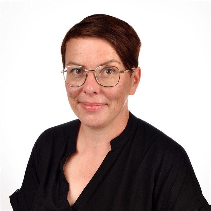 Henkilö, jolla on silmälasit ja musta toppi, hymyilee kameralle. Heidän hiuksensa ovat lyhyet ja tummat, ja tausta on pelkkä valkoinen.