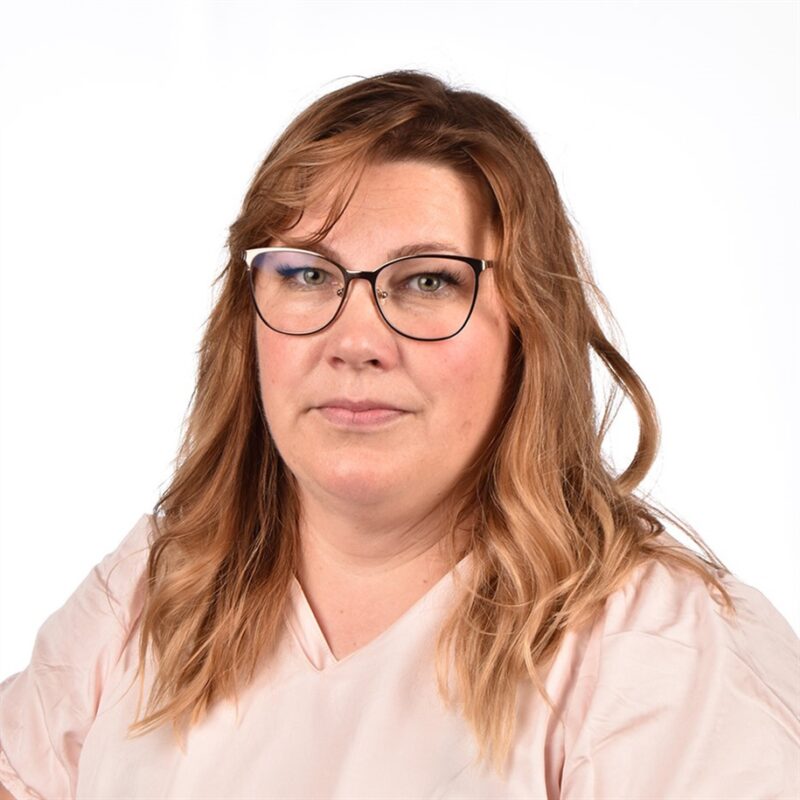 Naisella, jolla on aaltoilevat, olkapäille ulottuvat vaaleanruskeat hiukset ja silmälasit, on yllään vaaleanpunainen toppi. Hän poseeraa valkoista taustaa vasten.