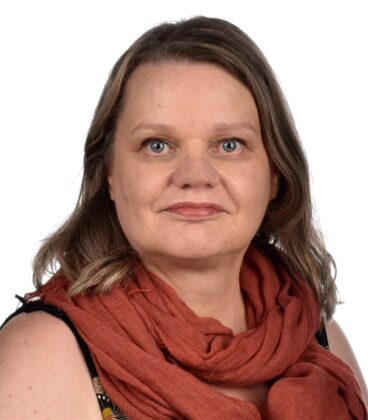 Olkapäille ulottuvat vaaleanruskeat hiukset omaava nainen, jolla on hihaton paita ja ruosteenvärinen huivi, katsoo suoraan kameraan tavallista valkoista taustaa vasten.