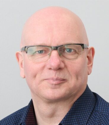 Keski-ikäinen kalju mies, jolla on silmälasit ja tummakuvioinen kauluspaita, katsoo suoraan kameraan neutraalilla ilmeellä vaaleanharmaata taustaa vasten.