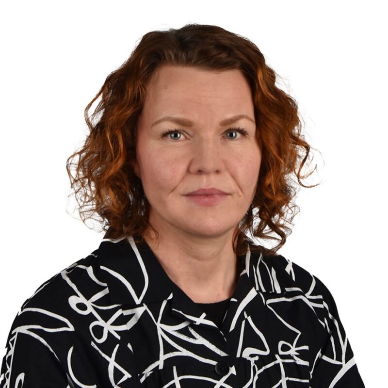 Nainen, jolla on aaltoilevat, olkapäille ulottuvat punaiset hiukset, yllään musta paita, jossa on valkoinen abstrakti kuviointi ja poseeraa pelkän valkoisen taustan edessä.