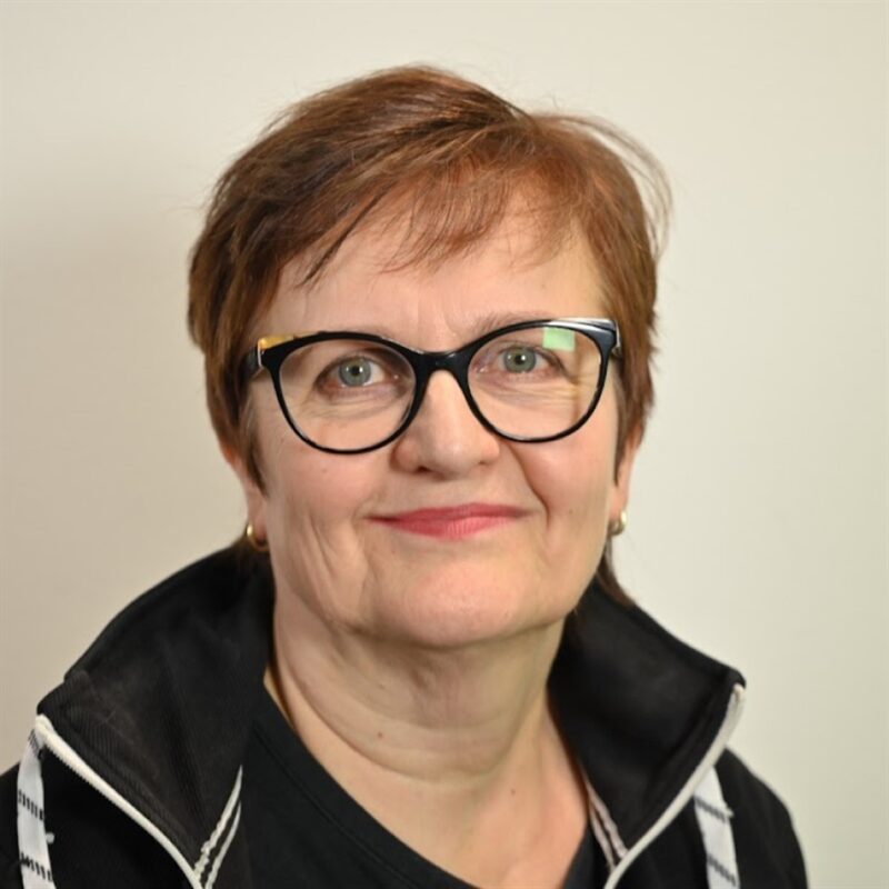 Nainen, jolla on lyhyet ruskeat hiukset ja silmälasit, hymyilee kameralle. Hänellä on yllään musta takki, jossa on korkea kaulus ja musta toppi. Tausta on tasainen ja vaalea.