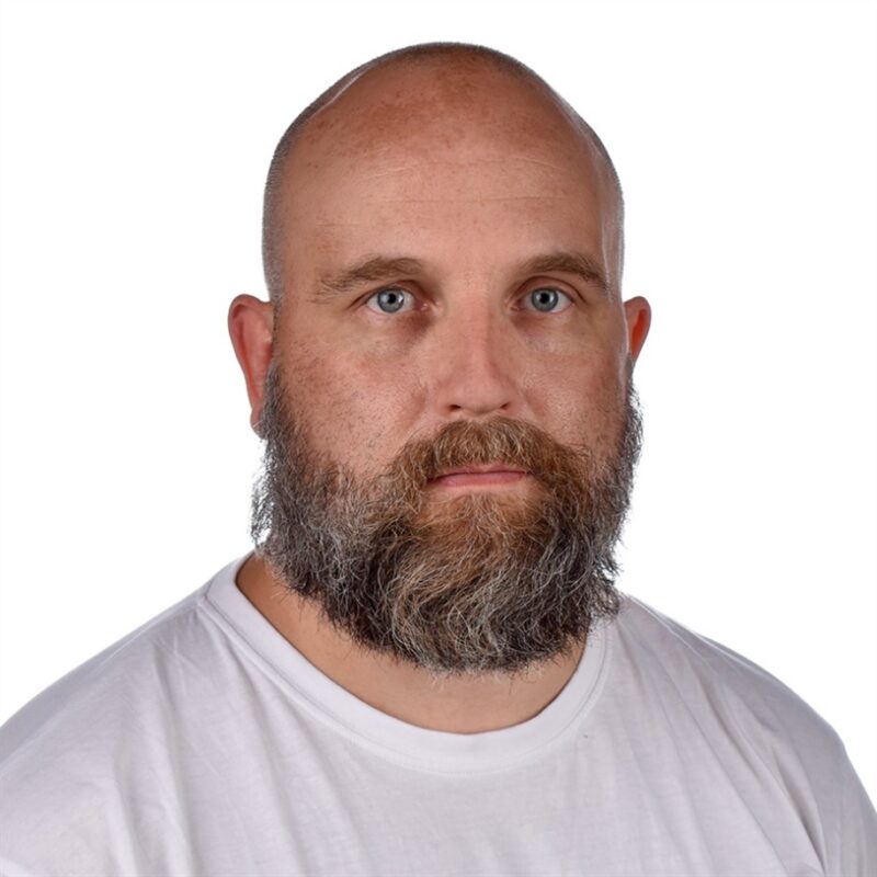 Parrakas mies, jolla on neutraali ilme, seisoo valkoista taustaa vasten. Hänellä on yllään valkoinen T-paita, hänellä on ajeltu pää ja hän katsoo suoraan kameraan.