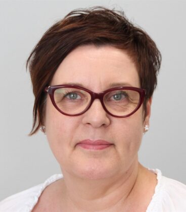 Lyhythiuksinen ruskeatukkainen ja tummanpunaisissa sankaisissa laseissa oleva nainen katsoo kameraan. Hänellä on valkoinen toppi ja pienet korvakorut, ja hän seisoo yksinkertaista vaaleanharmaata taustaa vasten.