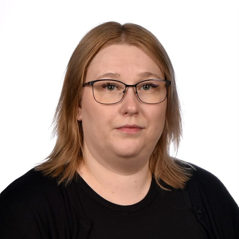 Muotokuva henkilöstä, jolla on vaaleanruskeat hiukset, silmälasit ja musta toppi, tavallista valkoista taustaa vasten. Niillä on neutraali ilme.