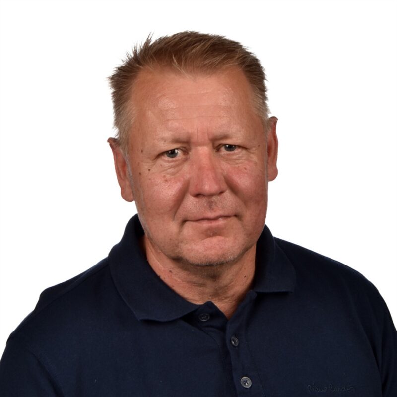 Vanhemmalla miehellä, jolla on lyhyet, vaaleat hiukset, on yllään tummansininen kauluspaita. Hän katsoo suoraan kameraan tavallista valkoista taustaa vasten.