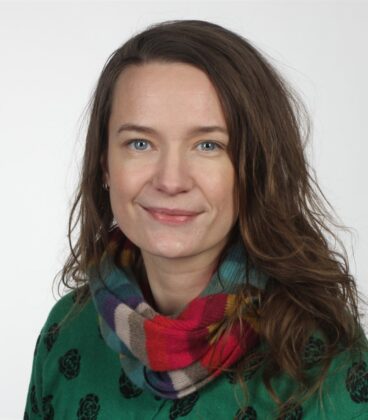 Pitkät ruskeat hiukset nainen hymyilee, yllään vihreä villapaita, jossa on musta kukkakuvio ja monivärinen huivi. Tausta on tavallinen valkoinen.